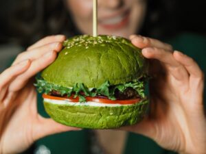 Burger tenu par deux main de chaque côté avec un pain vert, des tomate, de la salade et du fromage apparent.