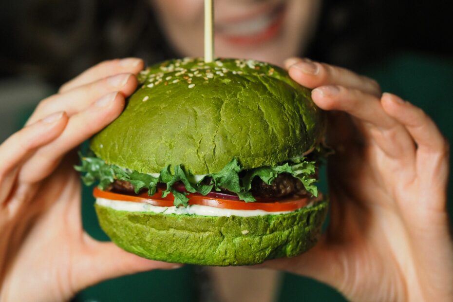 Burger tenu par deux main de chaque côté avec un pain vert, des tomate, de la salade et du fromage apparent.
